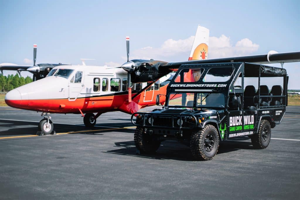 A white passenger tour airplane and a black custom Buck Wild ground tour vehicle parked side-by-side on an airport tarmac