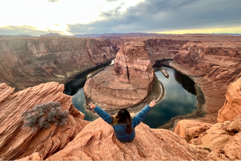 Person with arms raised enjoying the panoramic view of Horseshoe Bend at Grand Canyon under cloudy skies.