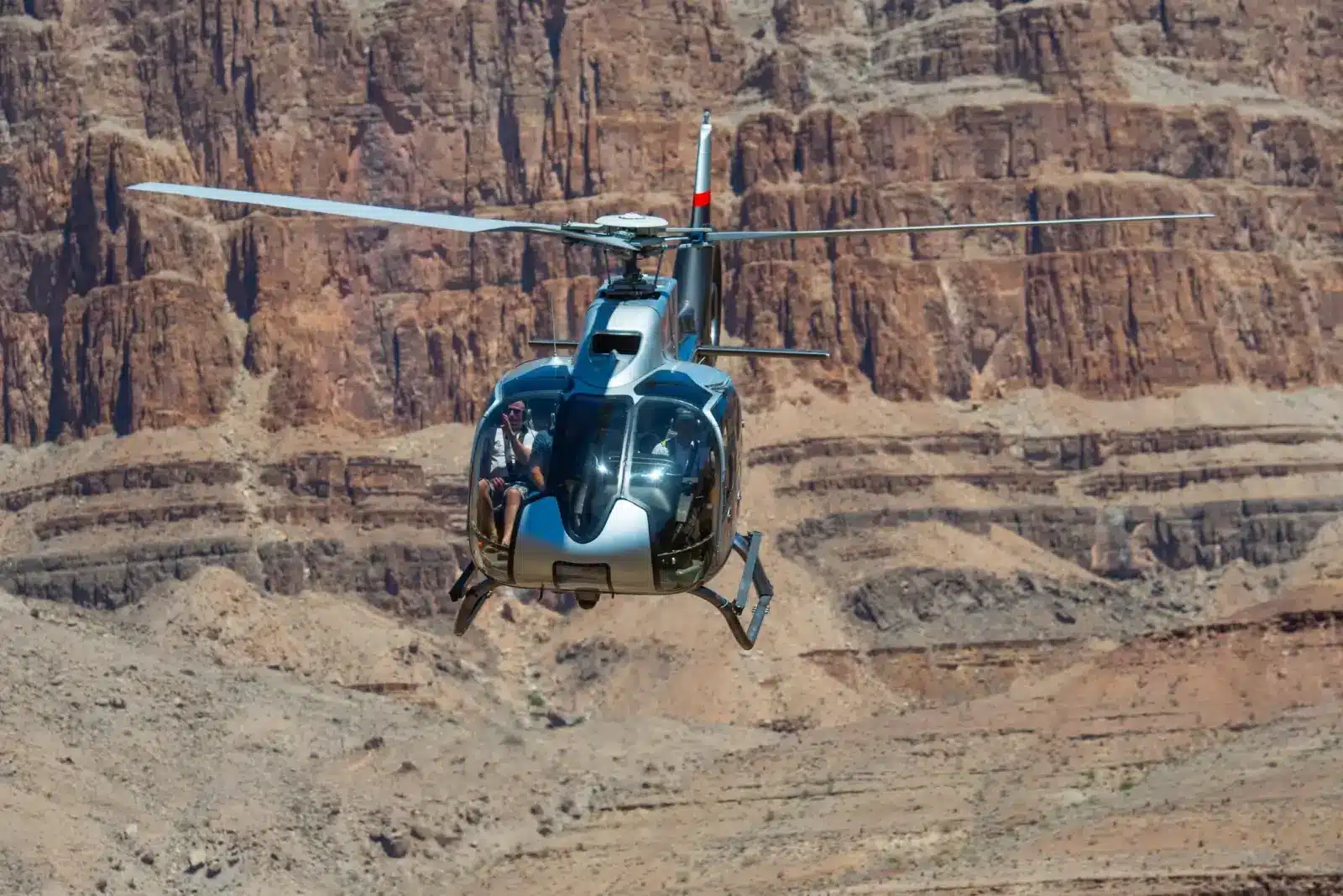 Silver and black tour helicopter flying low against the rugged rock walls of the Grand Canyon.